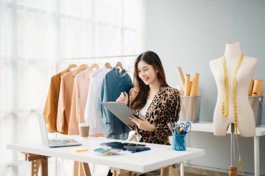 Asyalı genç kadın moda tasarımcılığının ofisinde ve elinde tablet, dizüstü bilgisayar ve akıllı telefon var.
