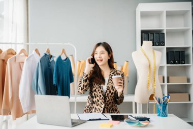 Asyalı genç kadın moda tasarımcılığının ofisinde ve elinde tablet, dizüstü bilgisayar ve akıllı telefon var.