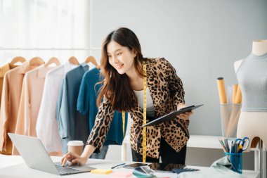 Asyalı genç kadın moda tasarımcılığının ofisinde ve elinde tablet, dizüstü bilgisayar ve akıllı telefon var.