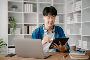 Erkek öğrenciler, Asyalı adam kütüphanesindeki kitaplardan dizüstü bilgisayar ve tablet kullanarak online bilgileri araştırıyorlar.