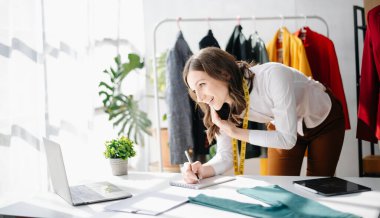 Moda tasarımcısı kadın akıllı telefonlardan konuşuyor ve stüdyosundaki dijital tablet bilgisayarlı dizüstü bilgisayarla raftaki kıyafetlerin yanında asılı duruyor.