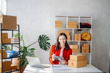 Startup small business SME, Entrepreneur owner woman using smartphone or tablet taking receive and checking online purchase shopping order to preparing pack product box. 