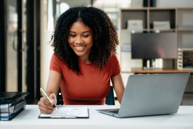 Tablet ve dizüstü bilgisayarda yazan genç bir kadın çalışan beyaz masada modern bir ofiste oturuyor.