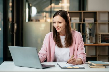 Kendine güvenen iş uzmanı, gülümseyen, dizüstü bilgisayarda yazan ve dijital tableti yaratıcı ofiste tutan çekici genç bir kadın.