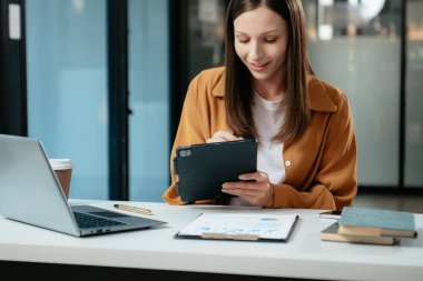 Kendine güvenen iş uzmanı, gülümseyen, dizüstü bilgisayarda yazan ve dijital tableti yaratıcı ofiste tutan çekici genç bir kadın.