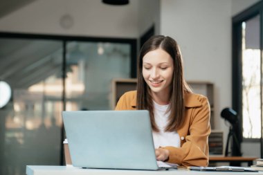 Kendine güvenen iş uzmanı, gülümseyen, dizüstü bilgisayarda yazan ve dijital tableti yaratıcı ofiste tutan çekici genç bir kadın.