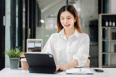 Çalışan not defteri, tablet ve laptop belgeleriyle modern ofiste çalışan Asyalı iş kadını.