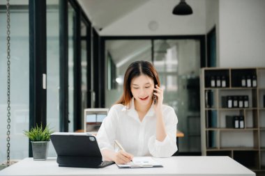Çalışan not defteri, tablet ve laptop belgeleriyle modern ofiste çalışan Asyalı iş kadını.