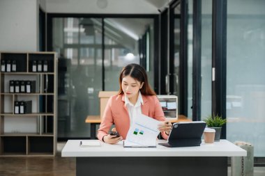 Çalışan not defteri, tablet ve laptop belgeleriyle modern ofiste çalışan Asyalı iş kadını.
