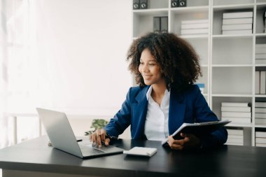 Tablet ve dizüstü bilgisayarda yazan genç, güzel Afrikalı kadın. Çalışan ahşap masada modern ofisin önünde oturuyor.
