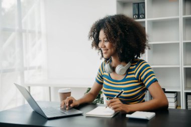 Üniversite kütüphanesinde okuyan çekici bir Afrikalı öğrenci, masada oturuyor, bilgisayar, tablet ve kulaklık kullanıyor ve görüntülü konuşma yapıyor.
