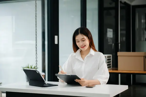 Çalışan not defteri, tablet ve laptop belgeleriyle modern ofiste çalışan Asyalı iş kadını.