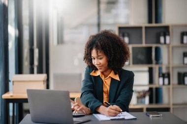Confident beautiful Asian businesswoman typing laptop computer and digital tablet while holding coffee at modern office