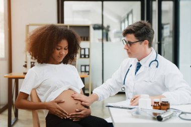 Hamile Afrikalı kadının klinikteki doktorla randevusu var. Stetoskop kullanan jinekolog erkek jinekolog kadın doğum uzmanı annesinin karnındaki bebeğin kalp atışlarını dinliyor. Hamilelik, sağlık konsepti