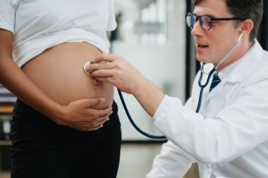 Hamile Afrikalı kadının klinikteki doktorla randevusu var. Stetoskop kullanan jinekolog erkek jinekolog kadın doğum uzmanı annesinin karnındaki bebeğin kalp atışlarını dinliyor. Hamilelik, sağlık konsepti