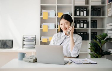 İş kadını Asyalı kadın telefonda konuşuyor ve ofiste otururken bilgisayarını gülümsüyor.