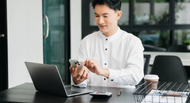 Masada oturan Asyalı bir adam dizüstü bilgisayar kullanarak ofiste Finans ve Pazarlama ile uğraşıyor.