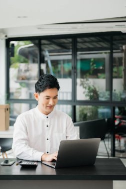 Masada oturan Asyalı bir adam dizüstü bilgisayar kullanarak ofiste Finans ve Pazarlama ile uğraşıyor.