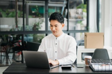 Masada oturan Asyalı bir adam dizüstü bilgisayar kullanarak ofiste Finans ve Pazarlama ile uğraşıyor.