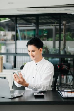 Masada oturan Asyalı bir adam dizüstü bilgisayar kullanarak ofiste Finans ve Pazarlama ile uğraşıyor.
