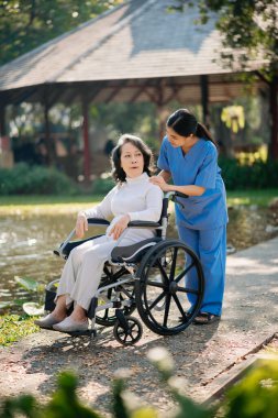 Asyalı yaşlı, yaşlı, yaşlı, tekerlekli sandalyede, Asyalı, dikkatli bir bakıcı ve bahçede yürüyen bir hastayı cesaretlendiren bir kadın. Bakıcının ve kıdemli sağlık sigortasının dikkatine.
