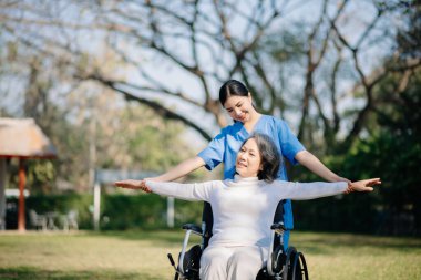 Asyalı yaşlı, yaşlı, yaşlı, tekerlekli sandalyede, Asyalı, dikkatli bir bakıcı ve bahçede yürüyen bir hastayı cesaretlendiren bir kadın. Bakıcının ve kıdemli sağlık sigortasının dikkatine.
