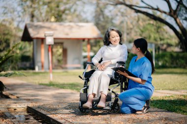Asyalı yaşlı, yaşlı, yaşlı, tekerlekli sandalyede, Asyalı, dikkatli bir bakıcı ve bahçede yürüyen bir hastayı cesaretlendiren bir kadın. Bakıcının ve kıdemli sağlık sigortasının dikkatine.