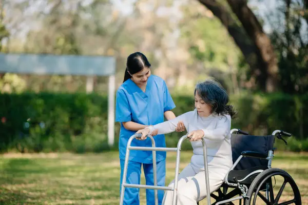 Asyalı genç bakıcı hemşire yaşlı bir erkeğin dışarıda yürümesini destekliyor. Uzman doktorlar yaşlı yetişkinlere yardım eder ve onlarla ilgilenir. Huzurevi hastane bahçesi konsepti