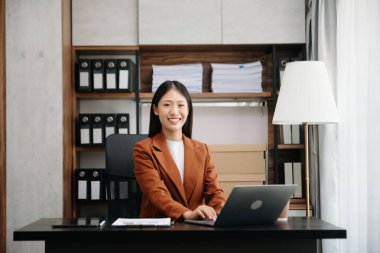 Genç Asyalı kadın gülerek not defteri, tablet ve dizüstü bilgisayarla yazmayı düşünüyor evden çalışıyor, ofisteki kameraya bakıyor.