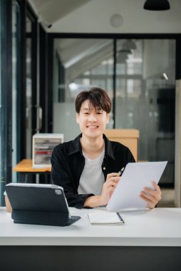 Modern ofiste elinde not defteri ve tabletle gülümseyen kendine güvenen Asyalı bir adam.