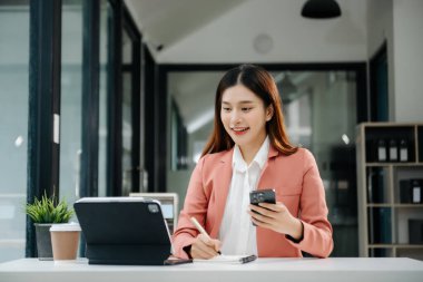 Çalışan not defteri, tablet ve laptop belgeleriyle modern ofiste çalışan Asyalı iş kadını.