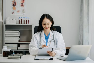 Beyaz üniformalı, kendine güvenen Asyalı kadın doktor masa başında bilgisayar üzerinde çalışıyor. Klinikteki tıp dergisine gülümseyen dizüstü bilgisayar yazıları.