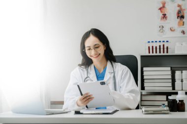 Beyaz üniformalı, kendine güvenen Asyalı kadın doktor masa başında bilgisayar üzerinde çalışıyor. Klinikteki tıp dergisine gülümseyen dizüstü bilgisayar yazıları.