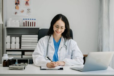 Beyaz üniformalı, kendine güvenen Asyalı kadın doktor masa başında bilgisayar üzerinde çalışıyor. Klinikteki tıp dergisine gülümseyen dizüstü bilgisayar yazıları.