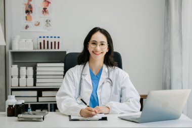 Beyaz üniformalı, kendine güvenen Asyalı kadın doktor masa başında bilgisayar üzerinde çalışıyor. Klinikteki tıp dergisine gülümseyen dizüstü bilgisayar yazıları.