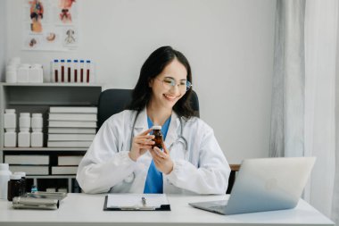 Beyaz üniformalı, kendine güvenen Asyalı kadın doktor masa başında bilgisayar üzerinde çalışıyor. Klinikteki tıp dergisine gülümseyen dizüstü bilgisayar yazıları.