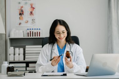 Beyaz üniformalı, kendine güvenen Asyalı kadın doktor masa başında bilgisayar üzerinde çalışıyor. Klinikteki tıp dergisine gülümseyen dizüstü bilgisayar yazıları.