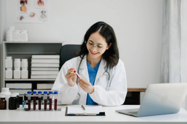 Beyaz üniformalı, kendine güvenen Asyalı kadın doktor masa başında bilgisayar üzerinde çalışıyor. Klinikteki tıp dergisine gülümseyen dizüstü bilgisayar yazıları.