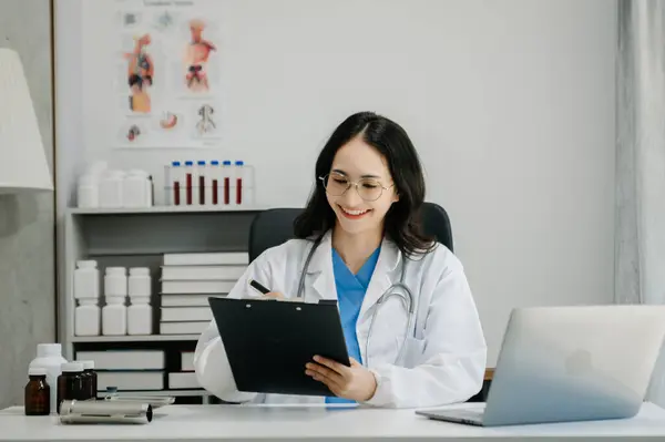Beyaz üniformalı, kendine güvenen Asyalı kadın doktor masa başında bilgisayar üzerinde çalışıyor. Klinikteki tıp dergisine gülümseyen dizüstü bilgisayar yazıları.