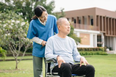 Asyalı yaşlı, yaşlı, yaşlı, tekerlekli sandalyede, Asyalı, dikkatli bir bakıcı ve bahçede yürüyen bir hastayı cesaretlendiren bir kadın. Bakıcının ve kıdemli sağlık sigortasının dikkatine.