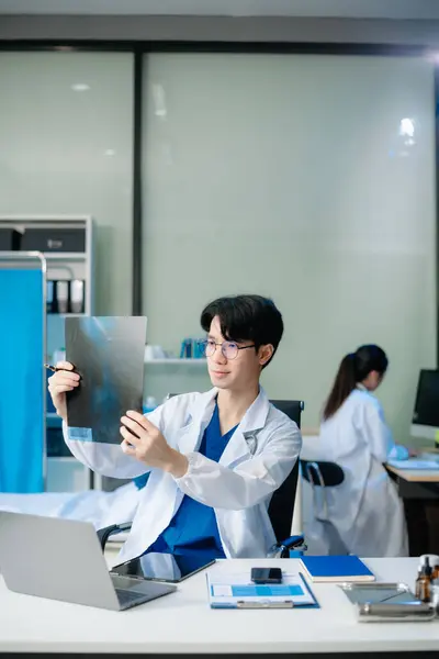 Confident young doctor in white medical uniform sit at desk working on computer. Smiling use laptop write in medical journal in clinic.