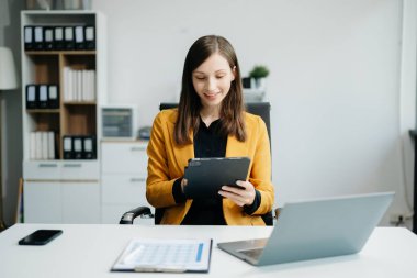 Kendine güvenen iş uzmanı, gülümseyen, dizüstü bilgisayarda yazan ve dijital tableti yaratıcı ofiste tutan çekici genç bir kadın.