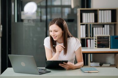 Güzel bir kadın çalışma yerinde otururken dizüstü bilgisayar ve tablet kullanıyor. İşine odaklan.