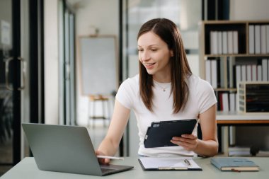 Güzel bir kadın çalışma yerinde otururken dizüstü bilgisayar ve tablet kullanıyor. İşine odaklan.