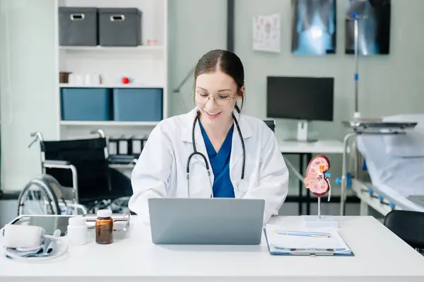 Confident young Caucasian female doctor in white medical uniform sit at desk working on computer. Smiling use laptop write in medical journal in clinic