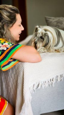 Renkli elbiseli bir kadın oturma odasında köpeğiyle sevgiyle etkileşime girer. Gülümseyen ve tüylü arkadaşını sevgiyle okşayan duygusal bağ açıktır..