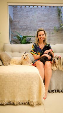 Gülümseyen bir kadın, etrafı iki köpek ve bir kedi dahil üç sevimli hayvanla çevrili süslü bir kanepede rahatça oturur. Köpeklerin kafasında fiyonklar var..