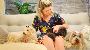 Gülümseyen bir kadın rahat bir koltukta oturur, yanında iki tane ışık kürklü köpek yavrusuyla siyah bir kediyi okşar. Sahne sıcak ve samimi bir aile atmosferini tasvir ediyor..