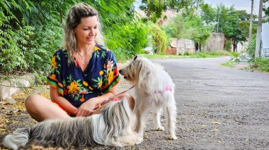 Gülümseyen, rahat bir kadın yemyeşil yemyeşil bir caddenin zemininde oturuyor. Sevecen bir şekilde iki sevimli köpekle iletişim kuruyor..