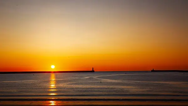 South Shields limanında gün doğumu, kuzey ve güney rıhtımlarında güneş doğuyor, yumuşak dalgalar üzerinde sarı bir parıltı yayıyor, Temmuz 2025